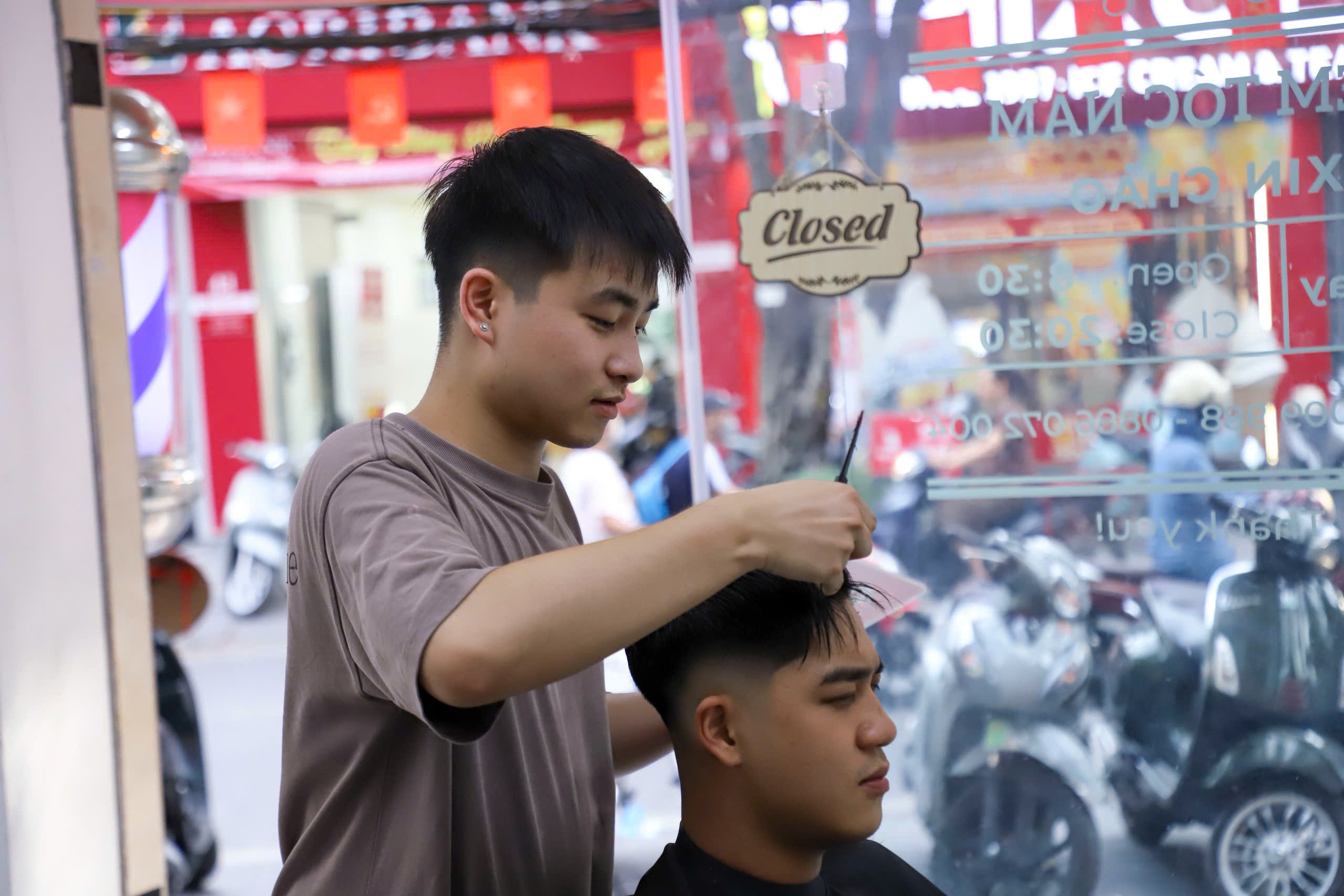 Nhóm bạn trẻ ngồi trước cửa hàng Tuyen Do Barbershop, phía trên là bảng hiệu và cột biển báo cắt tóc